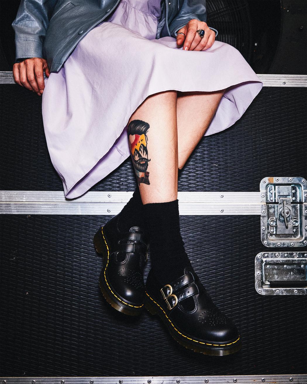 A model wearing a black leather Dr. Martens mary jane shoe with brass buckles.
